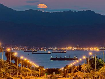 Sunset over a calm harbor with ships and city lights.