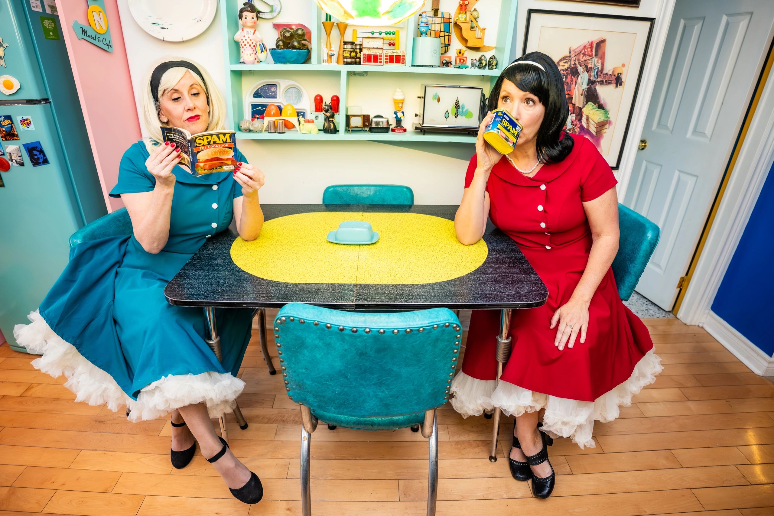Two women in retro dresses enjoying Spam-themed items in a colorful vintage kitchen.