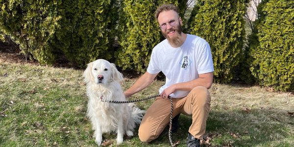 Man kneeling next to a white dog on grass with trees in the background.