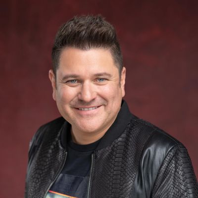 Smiling man in a textured black jacket against a dark red backdrop.