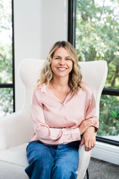 Smiling woman in a pink blouse sitting in a white chair by the window.