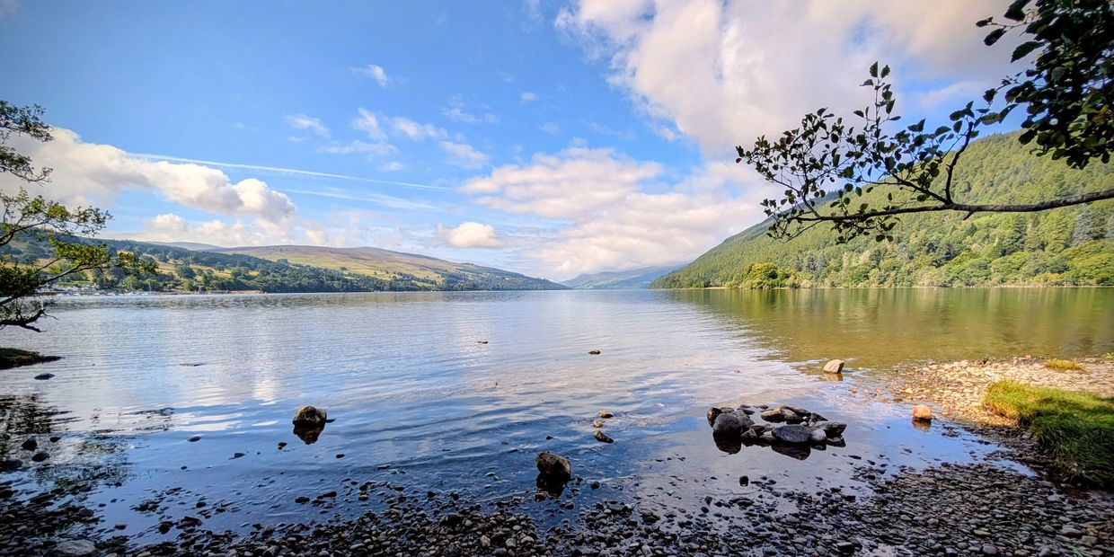 Calm lake with pebbled shore and trees under a blue sky

Loch Tay - Kenmore, Scotland.