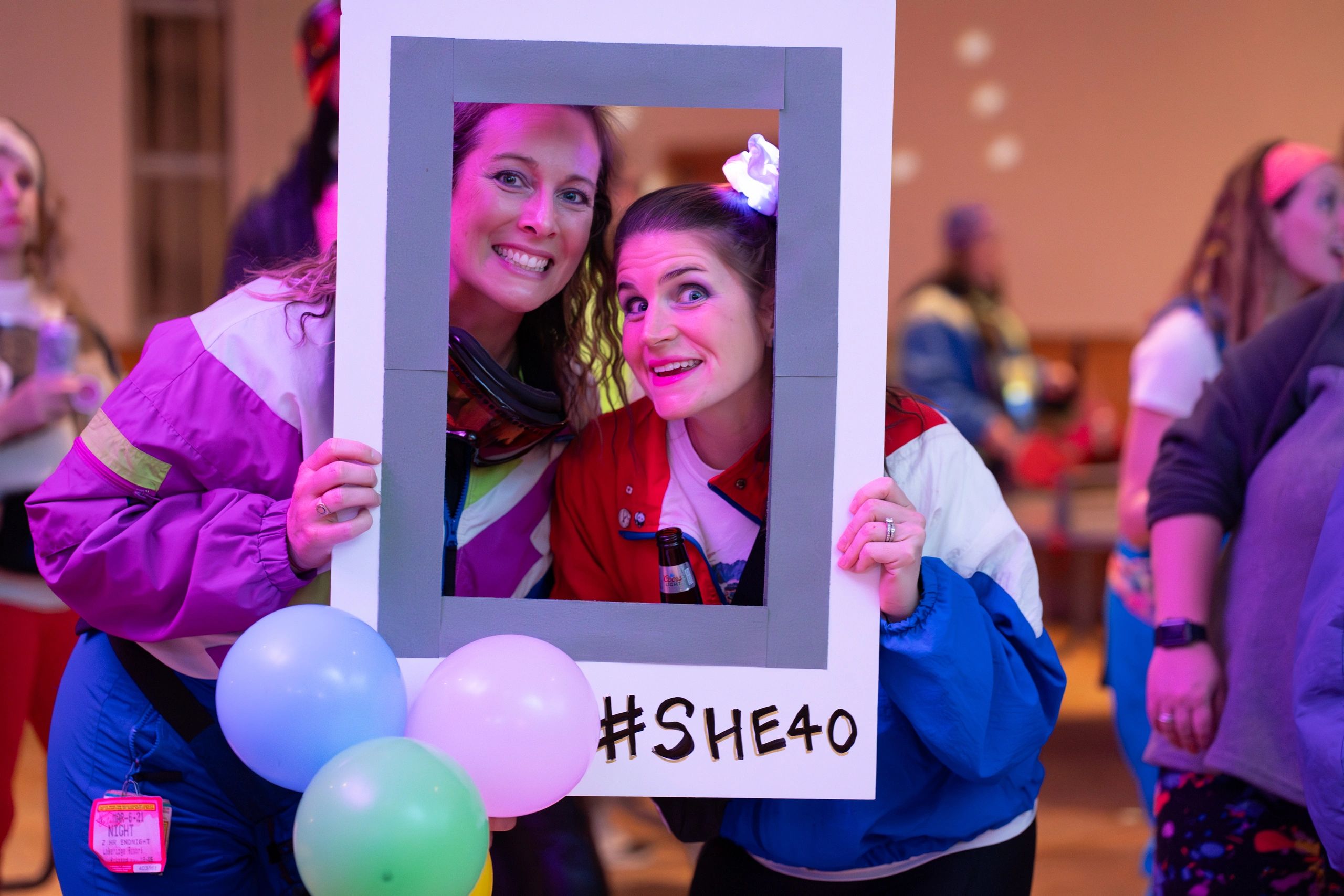 Two women smiling through a photo frame with #SHE40 and balloons.