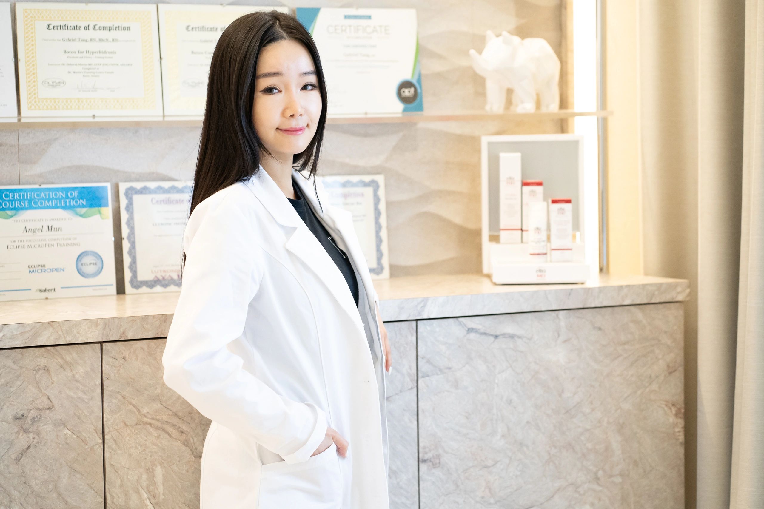 Confident female professional in a white coat standing in front of certificates.