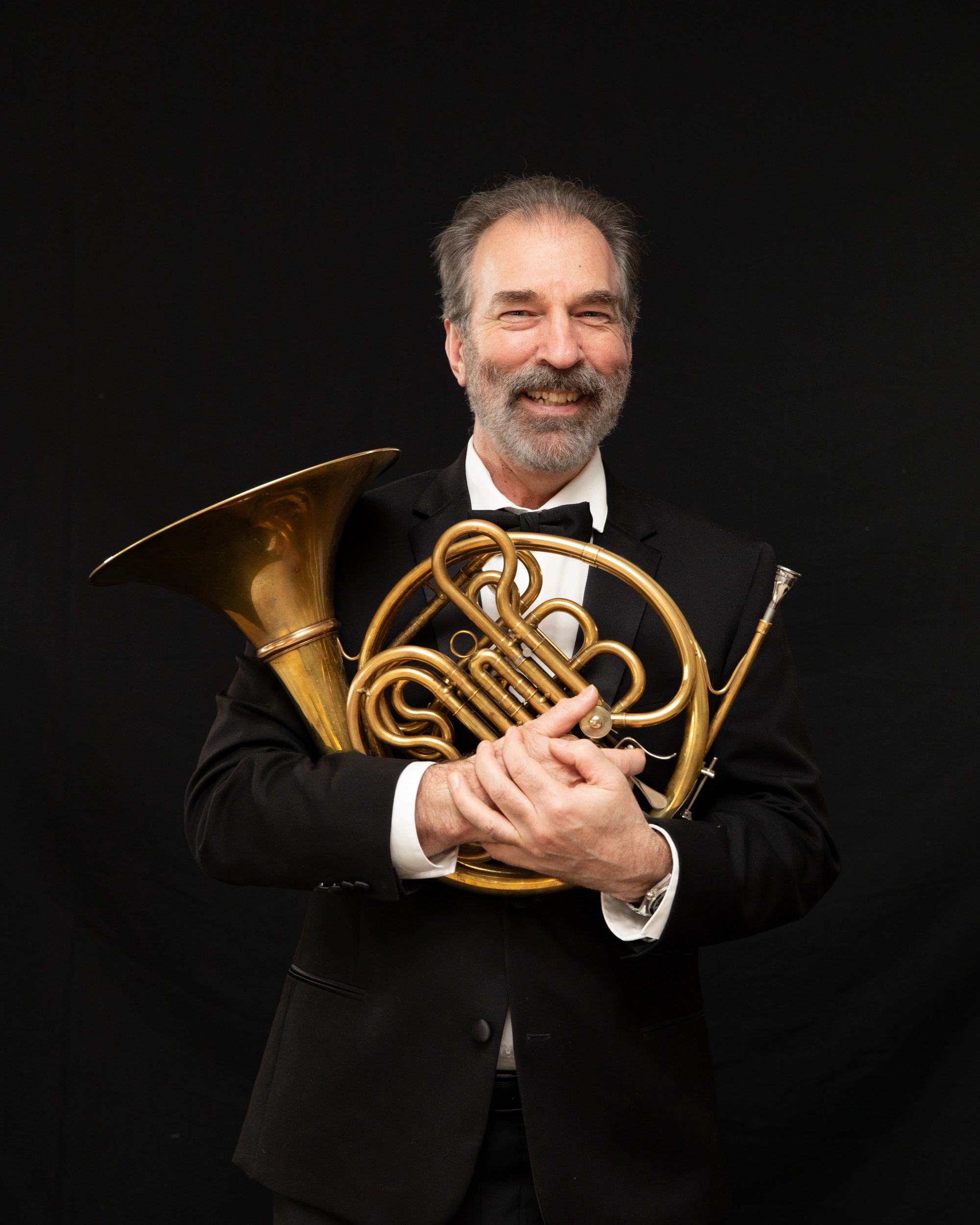 Smiling man in tuxedo holding a French horn against a black background.