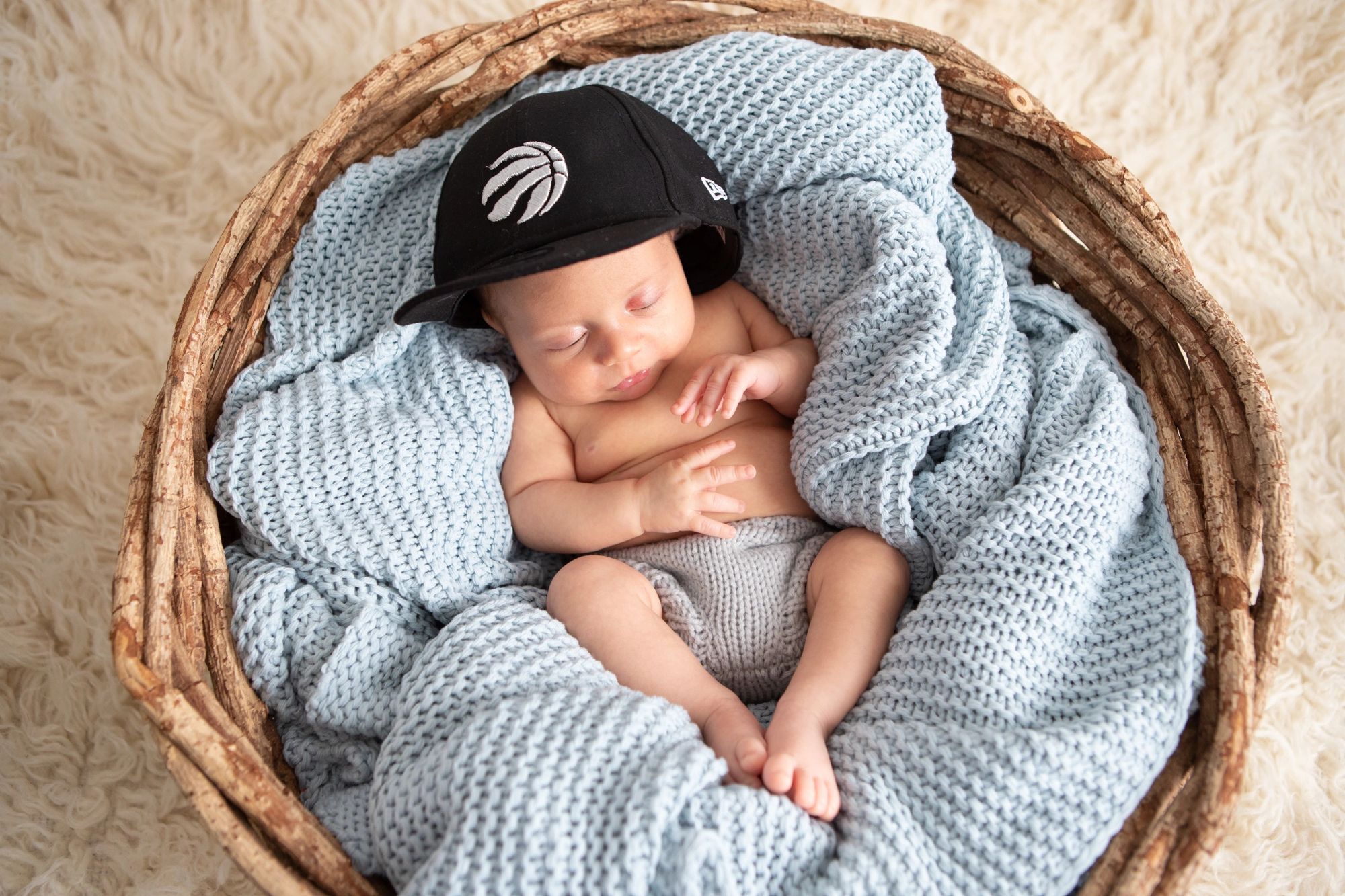 Sleeping baby wearing a black cap, wrapped in a blue knitted blanket inside a basket.