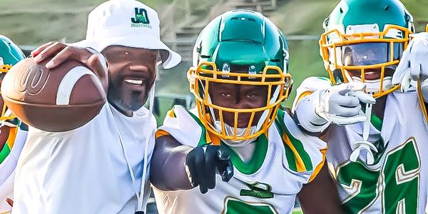 Coach and football players in green and white uniforms pointing at the camera on the field.
