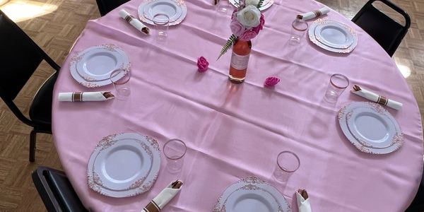 A round table with pink tablecloth set for six with elegant plates, napkins, and a floral centerpiece.