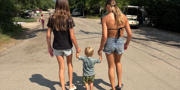 Two girls holding a toddler's hands walking down a road.