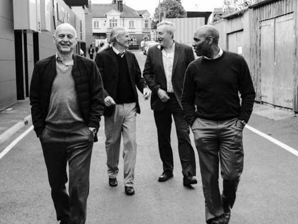 Four men casually walking and smiling down a street in a black and white photo.