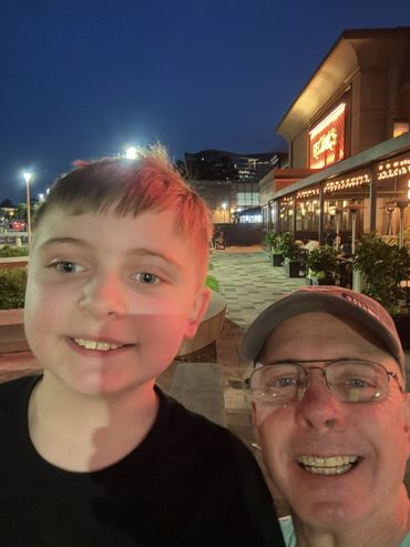 A smiling boy and man take a selfie outside a lit restaurant at night.