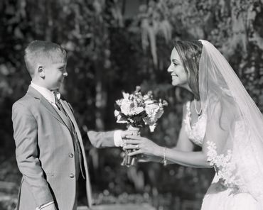 Bride smiling while receiving flowers from a young boy outdoors.