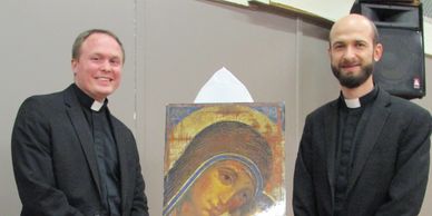 Two priests standing beside a religious painting of the Virgin Mary.