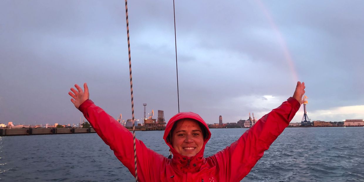 Person in red jacket raising arms with a rainbow over water and cityscape.
