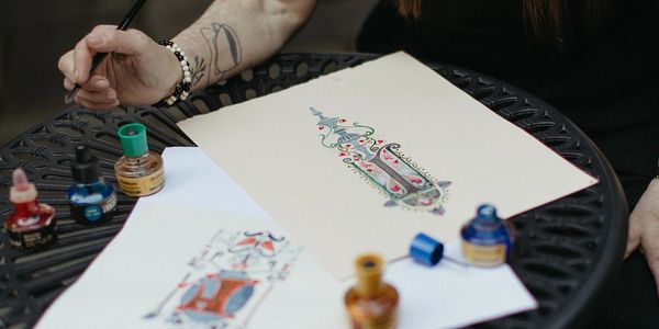 Person painting detailed designs using ink on paper at a table.