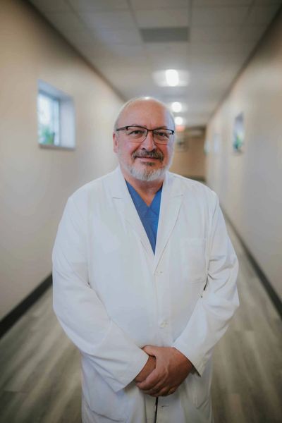 Older male doctor in white coat and glasses standing in hallway.
