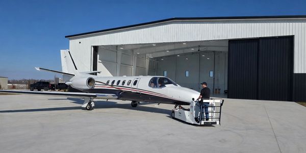 A person operating a tug vehicle to move a small private jet outside a hangar.