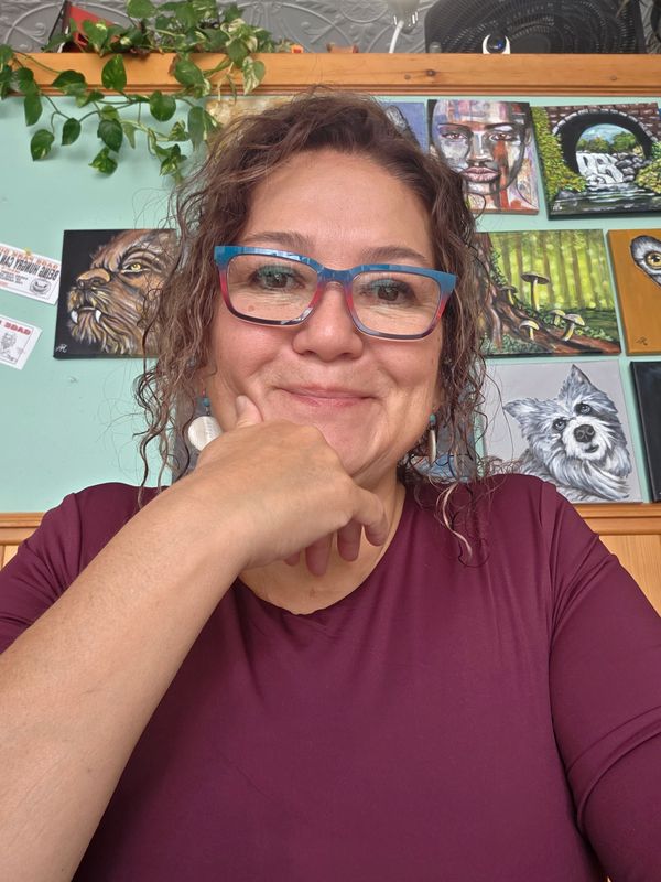 Woman with glasses smiling in front of colorful paintings indoors.