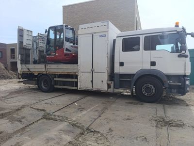 A white flatbed truck carrying a small excavator at a construction site.