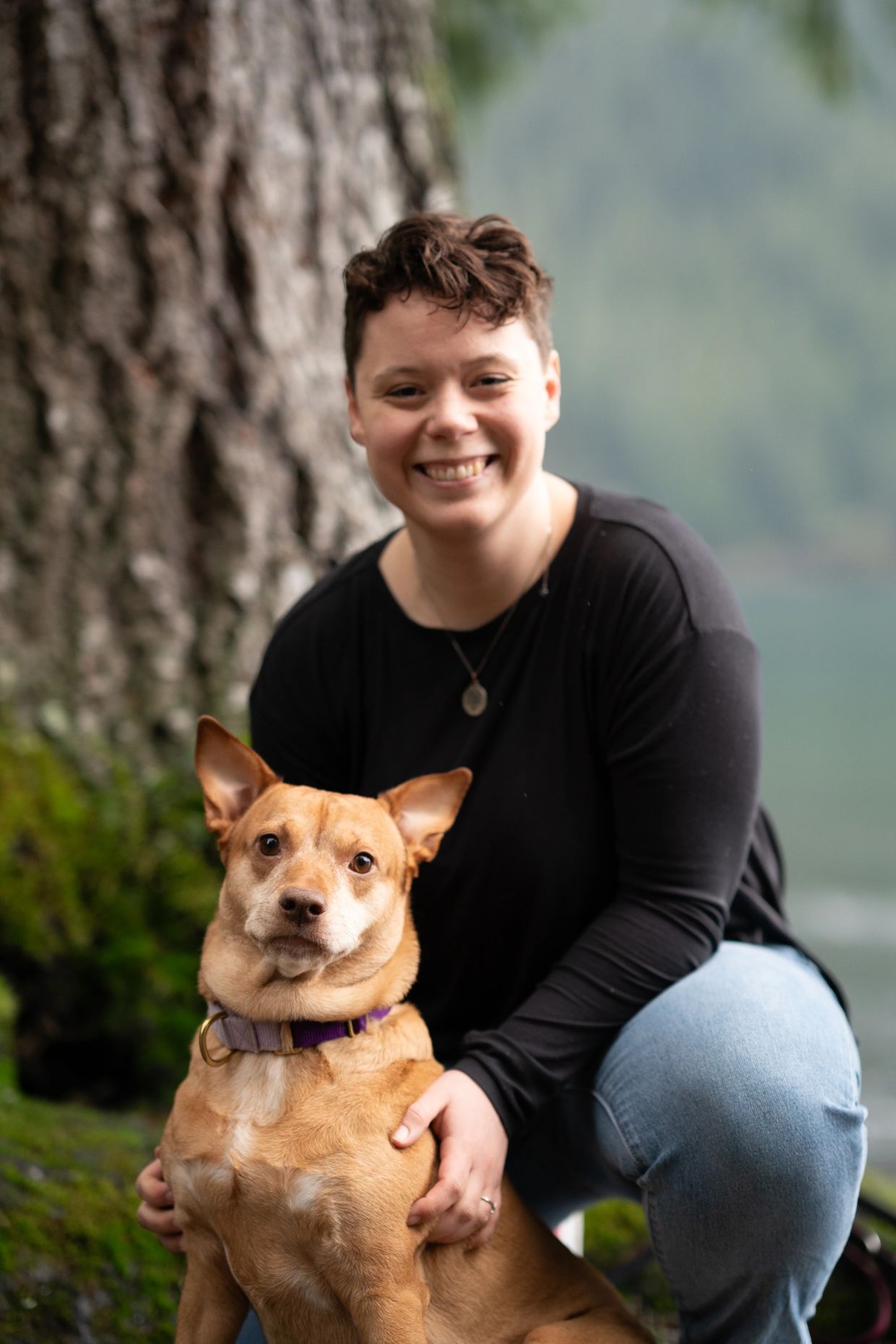 Person smiling with their dog outdoors by a tree.