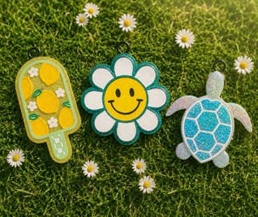 Three colorful keychains on grass with daisies: popsicle, smiley flower, and turtle.