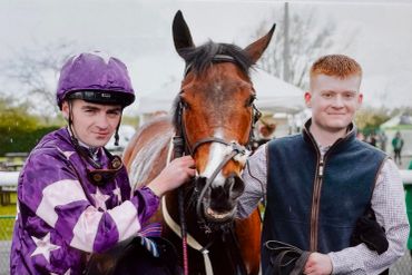 Two men posing with a horse, one dressed as a jockey in purple.