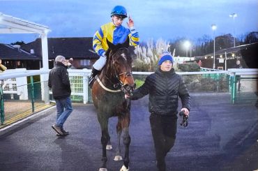 Jockey in yellow and blue outfit on horse led by a man in winter wear.