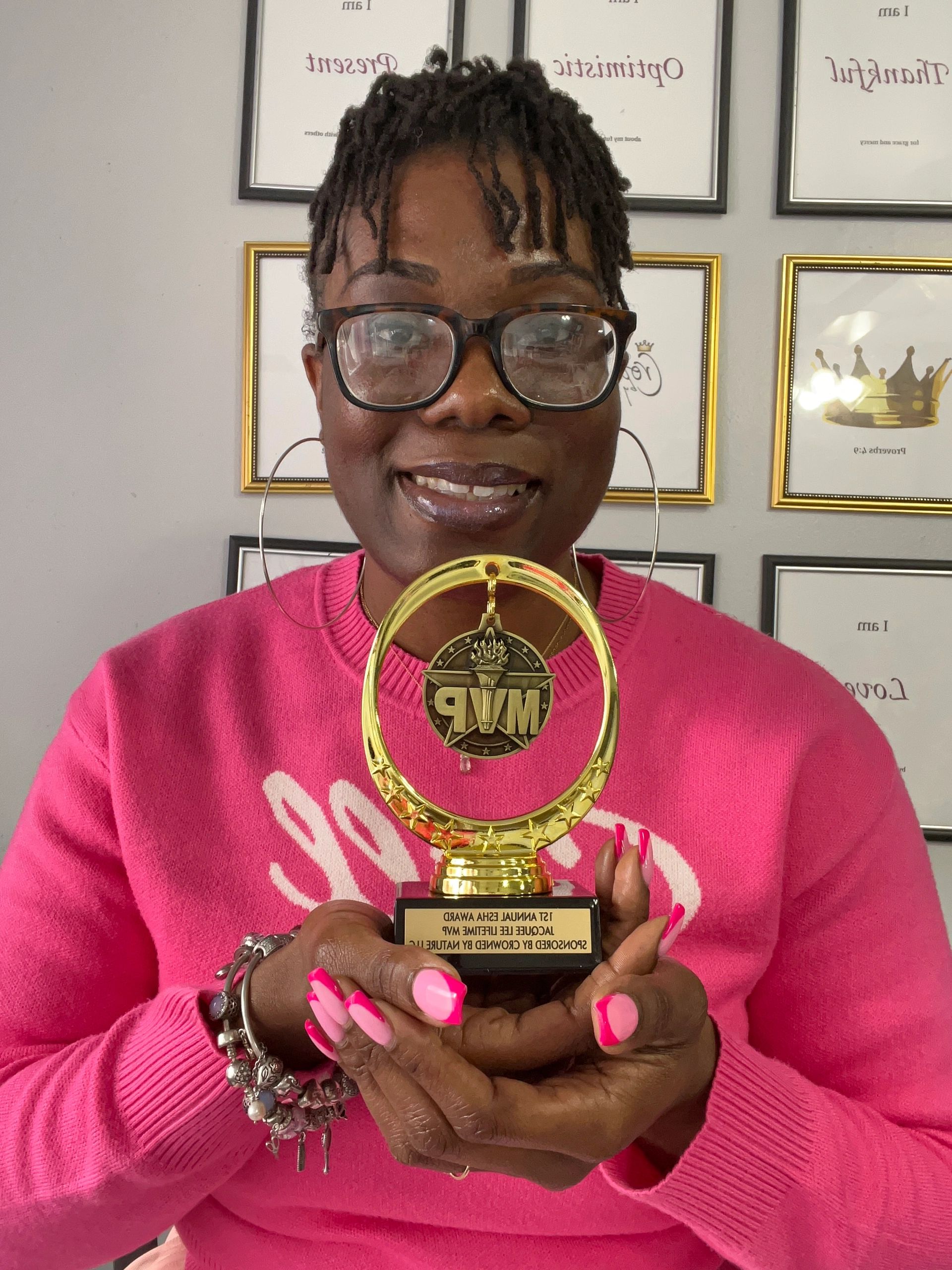A smiling Black woman with locs and glasses, wearing a pink sweater and holding a gold MVP trophy. 