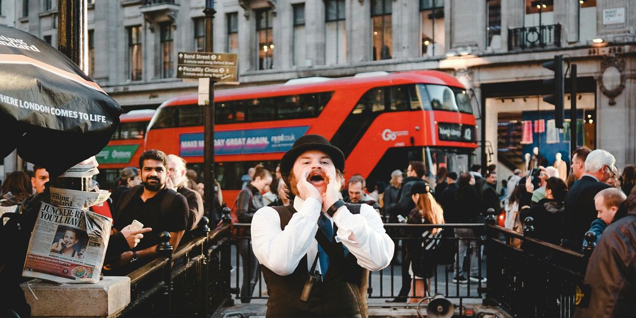A street performer shouts near a London Underground station with a red double-decker bus in the background.