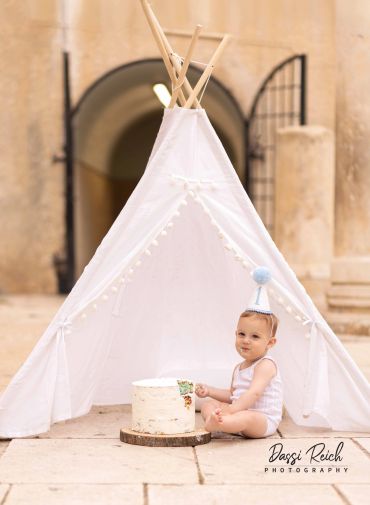 Baby in party hat sitting beside a white cake in front of a teepee.