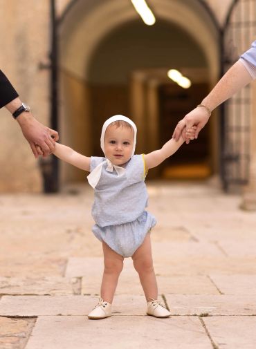 Baby holding hands with adults, standing on stone pavement.