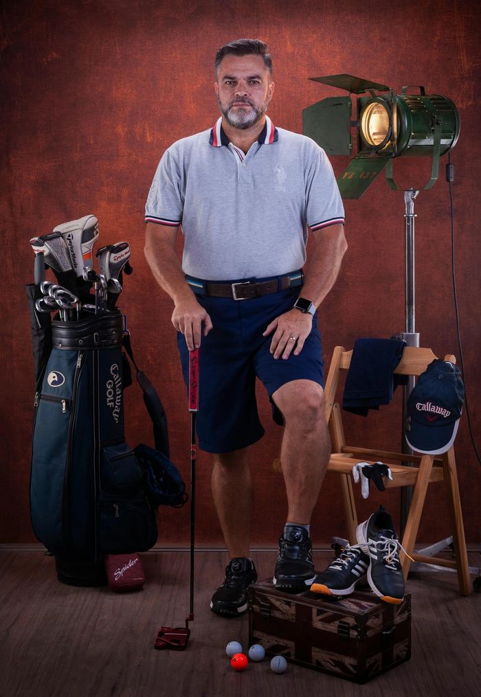 Man posing with golf equipment and a putter in a studio setting.
