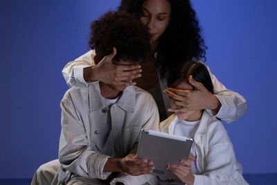A woman playfully covers two children's eyes as they look at a tablet.