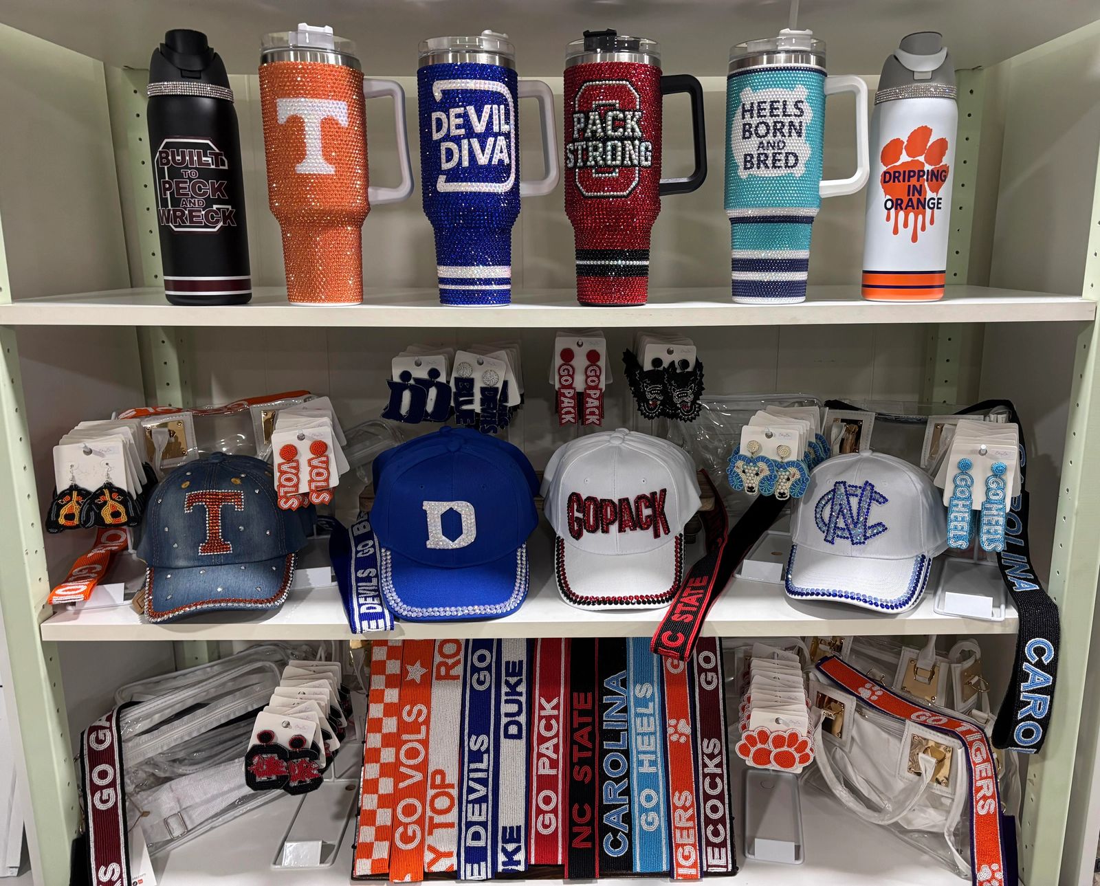 Rhinestone collegiate tumblers, denim caps, and earrings for Duke, UNC, NC State, Tennessee, and Clemson on display at BlingBox