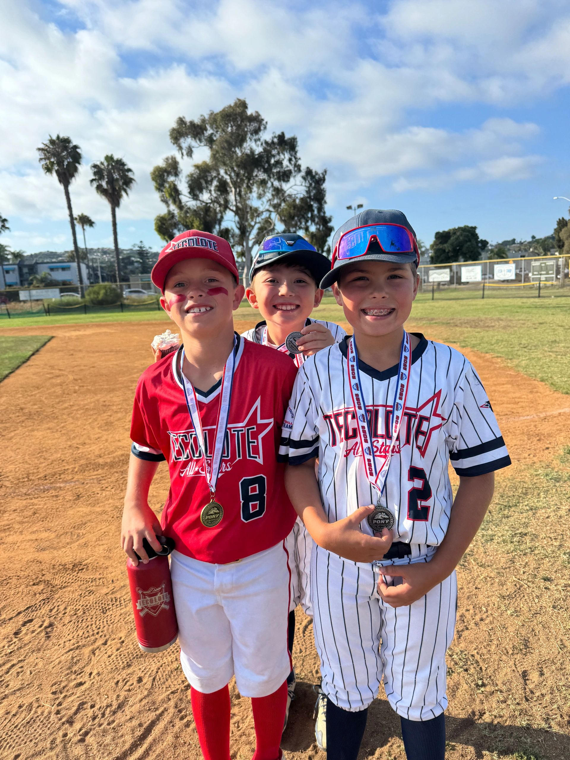 Three young baseball players proudly display their medals on a sunny field.