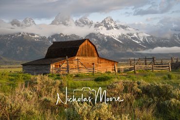 Rustic barn glowing in sunlight with snowy mountains and clouds in the background.