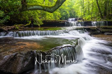 Multi-tiered waterfalls flow gently through a lush green forest.