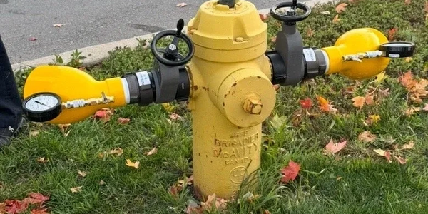 Yellow fire hydrant with attached pressure gauges and valves on a grassy roadside.