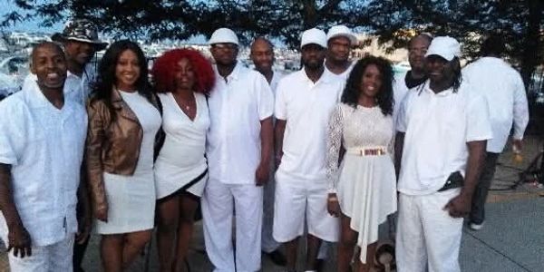 Group of people dressed mostly in white, posing outdoors under trees.