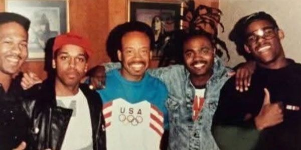 Five men posing together indoors, one wearing a USA Olympic sweater.