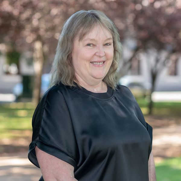 Smiling woman in black shirt outdoors with blurred trees and buildings behind.