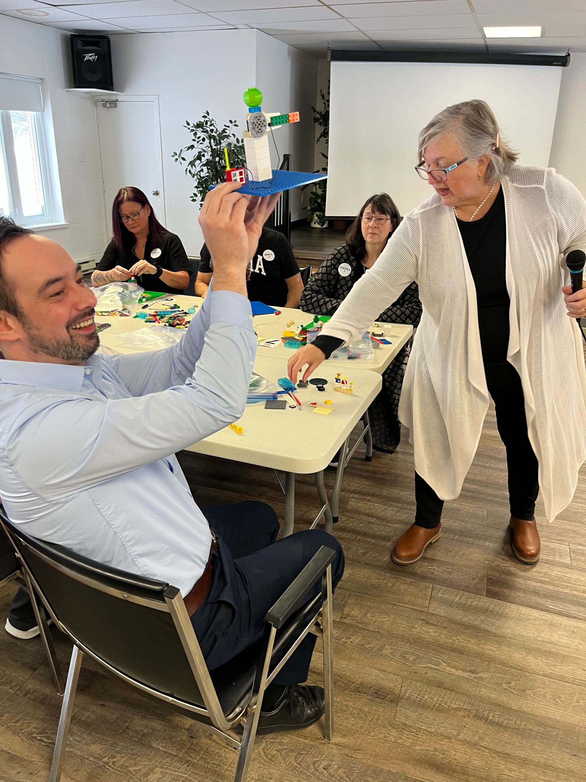 Man proudly shows a LEGO creation as a woman speaks to a group.