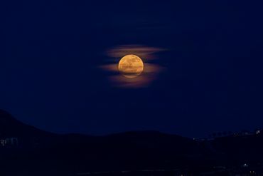 Full moon glowing through thin clouds over dark hills at night.