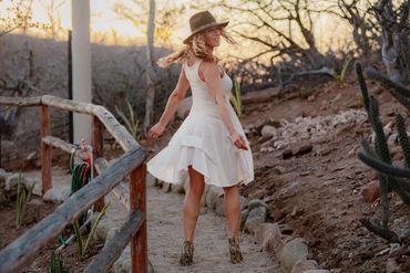 Woman in white dress and hat walking on rustic path at sunset.