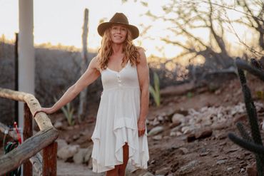 Woman in white dress and hat stands outdoors at sunset.
