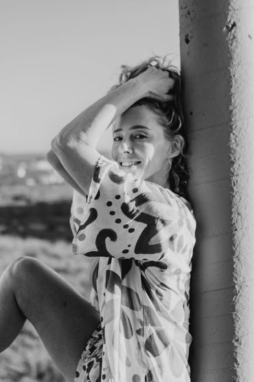 Young woman smiling, leaning against a pillar outdoors in patterned clothing.