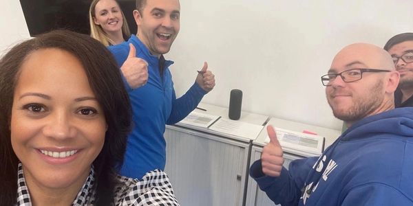 A group of five people smiling and giving thumbs up indoors.