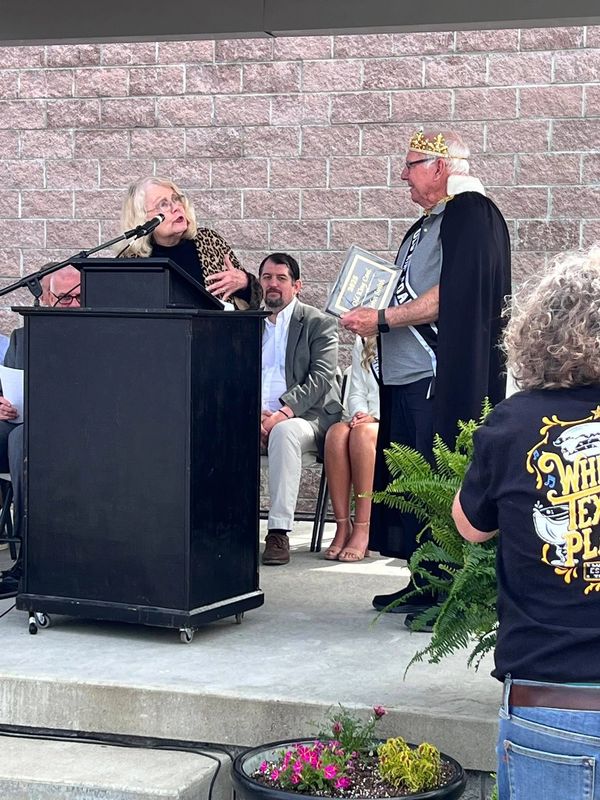 Man wearing a crown and cape receives an award from a woman at a podium in an outdoor event.