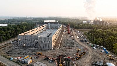 A large industrial building under construction with surrounding machinery and forest.