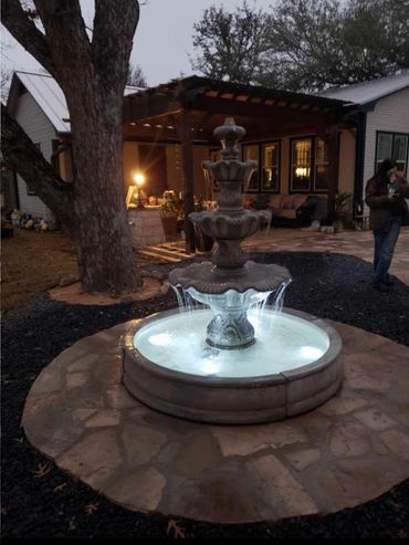 Illuminated three-tier stone fountain in a backyard at dusk.